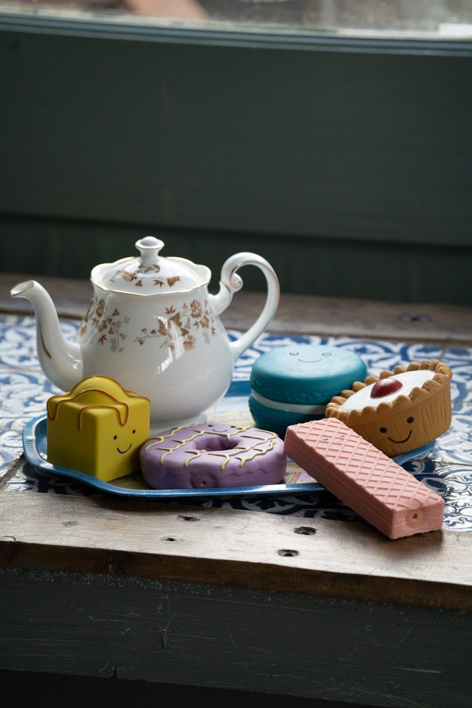 Teapot with colorful dog toys shaped like a cake, party ring, macaron, and cherry pie on a wooden surface.